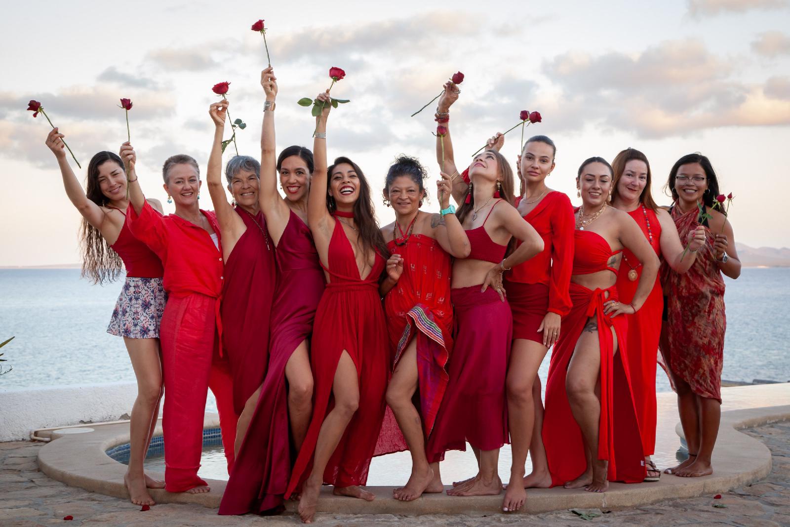 Un grupo de mujeres vestidas de rojo levanta rosas rojas junto a una alberca frente al mar, bajo un cielo nublado de atardecer.