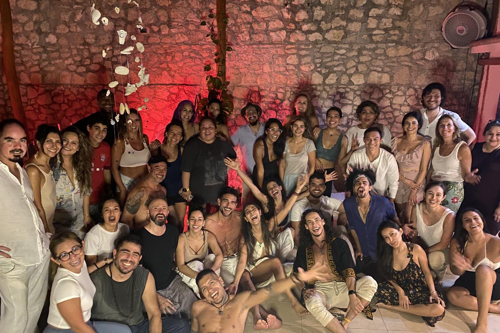 Foto de grupo nocturna de unas cuarenta personas sonrientes ante un muro de piedra iluminado en rojo, con conchas colgando del techo.