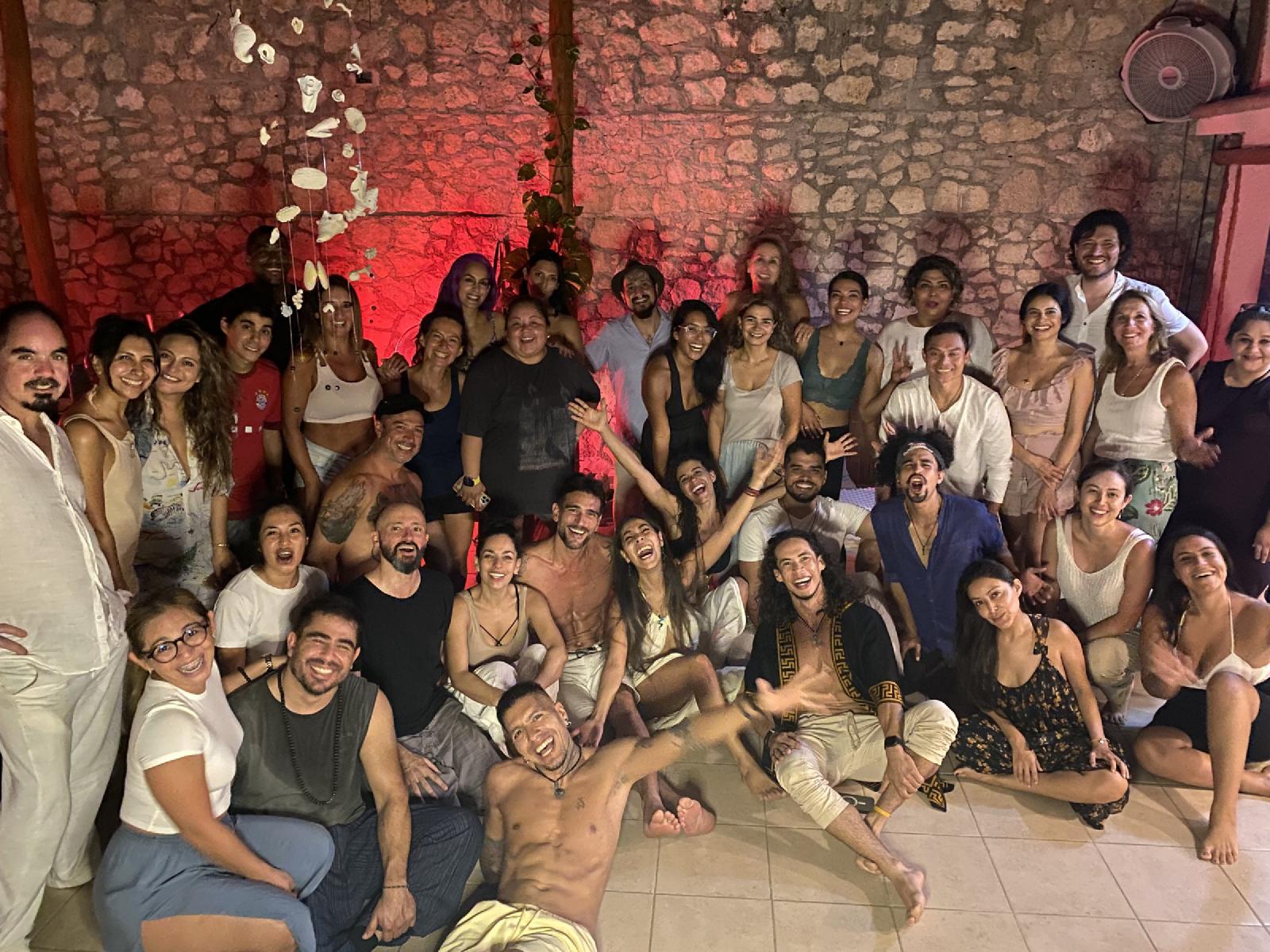 Foto de grupo nocturna de unas cuarenta personas sonrientes ante un muro de piedra iluminado en rojo, con conchas colgando del techo.