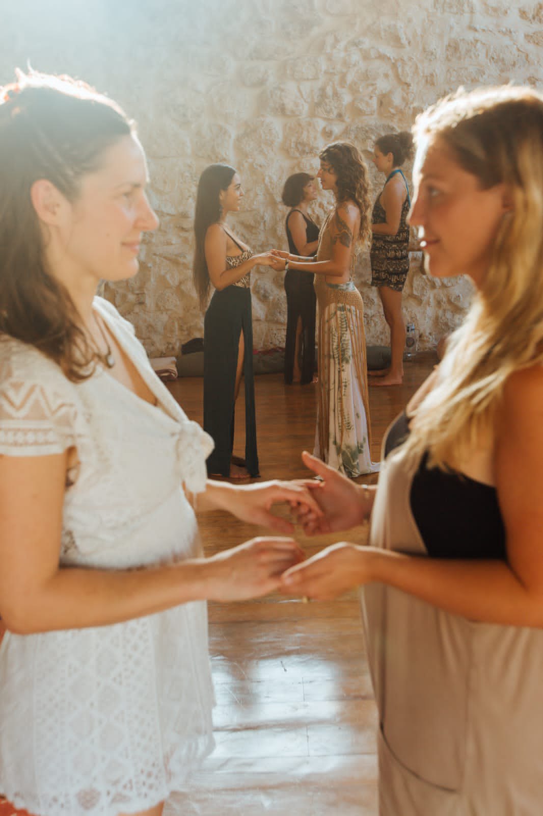 Dos mujeres de pie frente a frente, tomadas de las manos, en primer plano; al fondo otras parejas hacen lo mismo junto a un muro de piedra soleado.