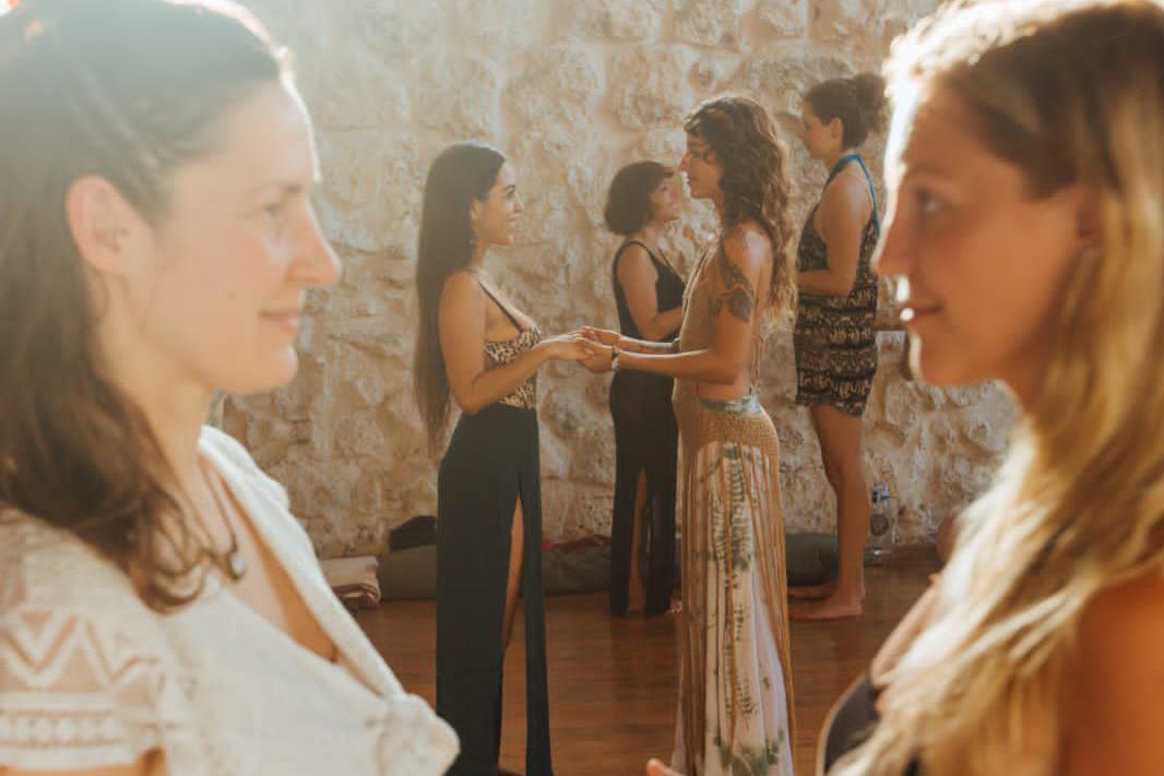 Dos mujeres de pie frente a frente, tomadas de las manos, en primer plano; al fondo otras parejas hacen lo mismo junto a un muro de piedra soleado.