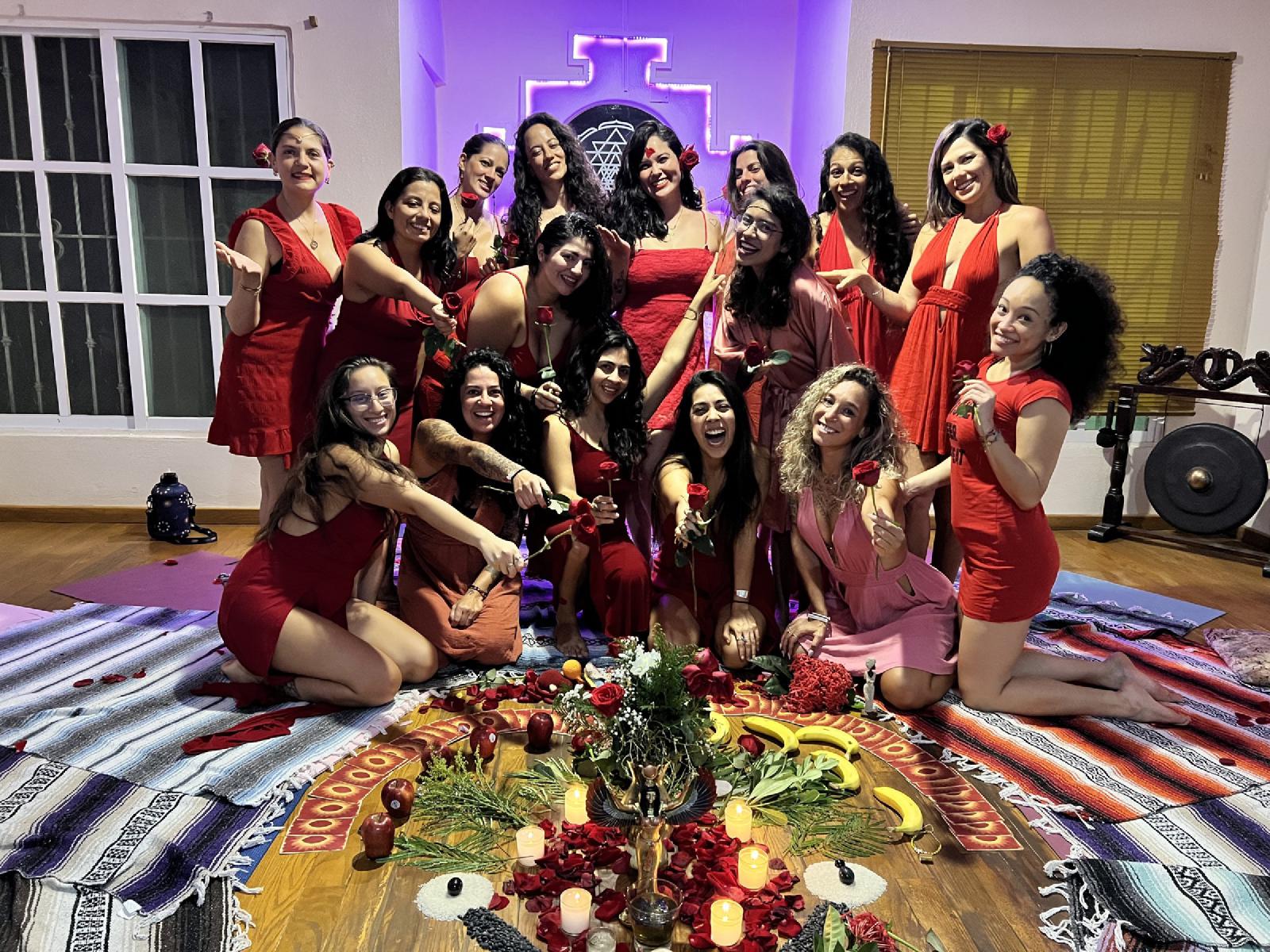 Mujeres vestidas de rojo sostienen rosas rojas, sonrientes, alrededor de un altar en el suelo con velas, pétalos, frutas y una figura de Isis alada.