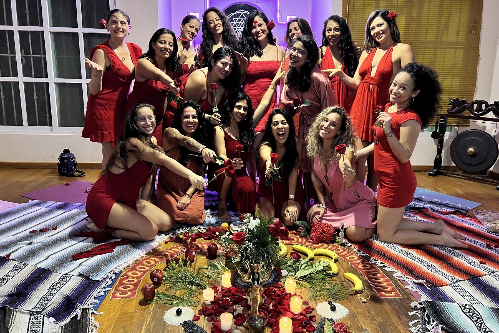 Mujeres vestidas de rojo sostienen rosas rojas, sonrientes, alrededor de un altar en el suelo con velas, pétalos, frutas y una figura de Isis alada.