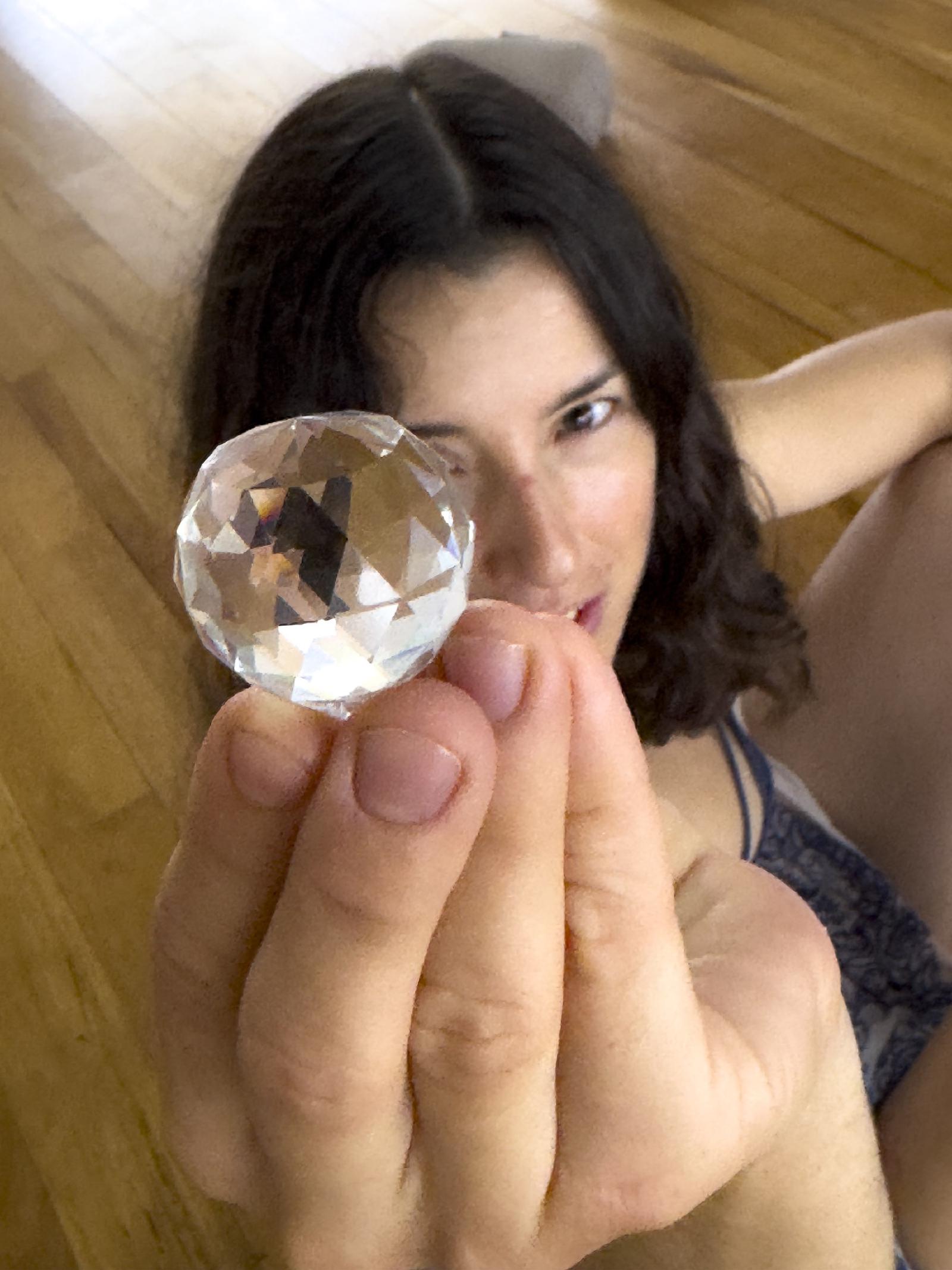 Una mujer recostada en un piso de madera sostiene una esfera de cristal tallado frente a su ojo, mirando a la cámara.