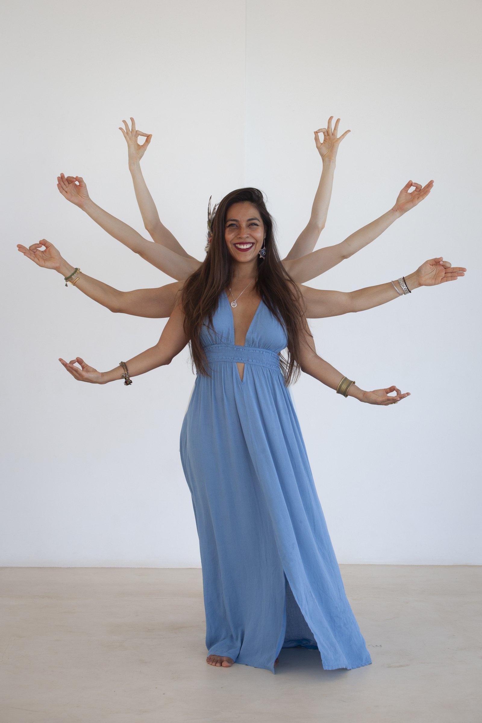 La Dra. Pamela Corona, con vestido largo azul, sonríe ante una pared blanca mientras varios brazos detrás de ella forman mudras, como una diosa de muchos brazos.