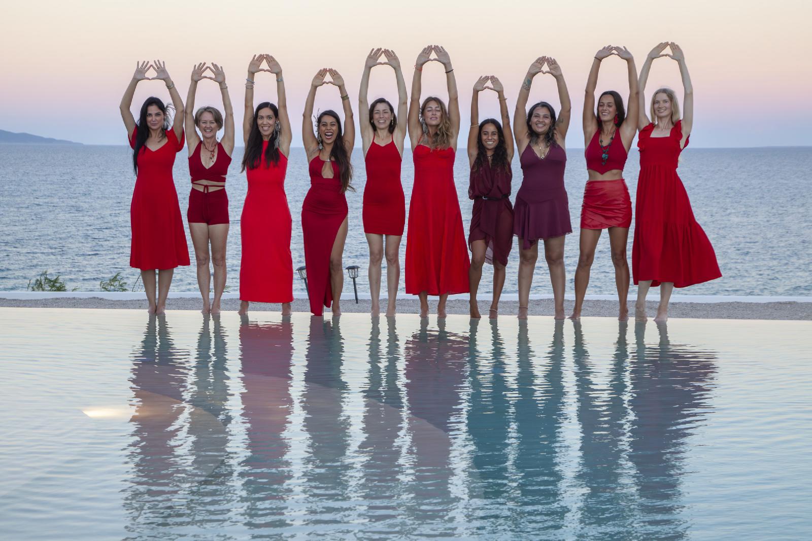 Diez mujeres vestidas de rojo, de pie en el borde de una alberca infinita al atardecer, forman un triángulo con las manos sobre la cabeza; el agua las refleja.