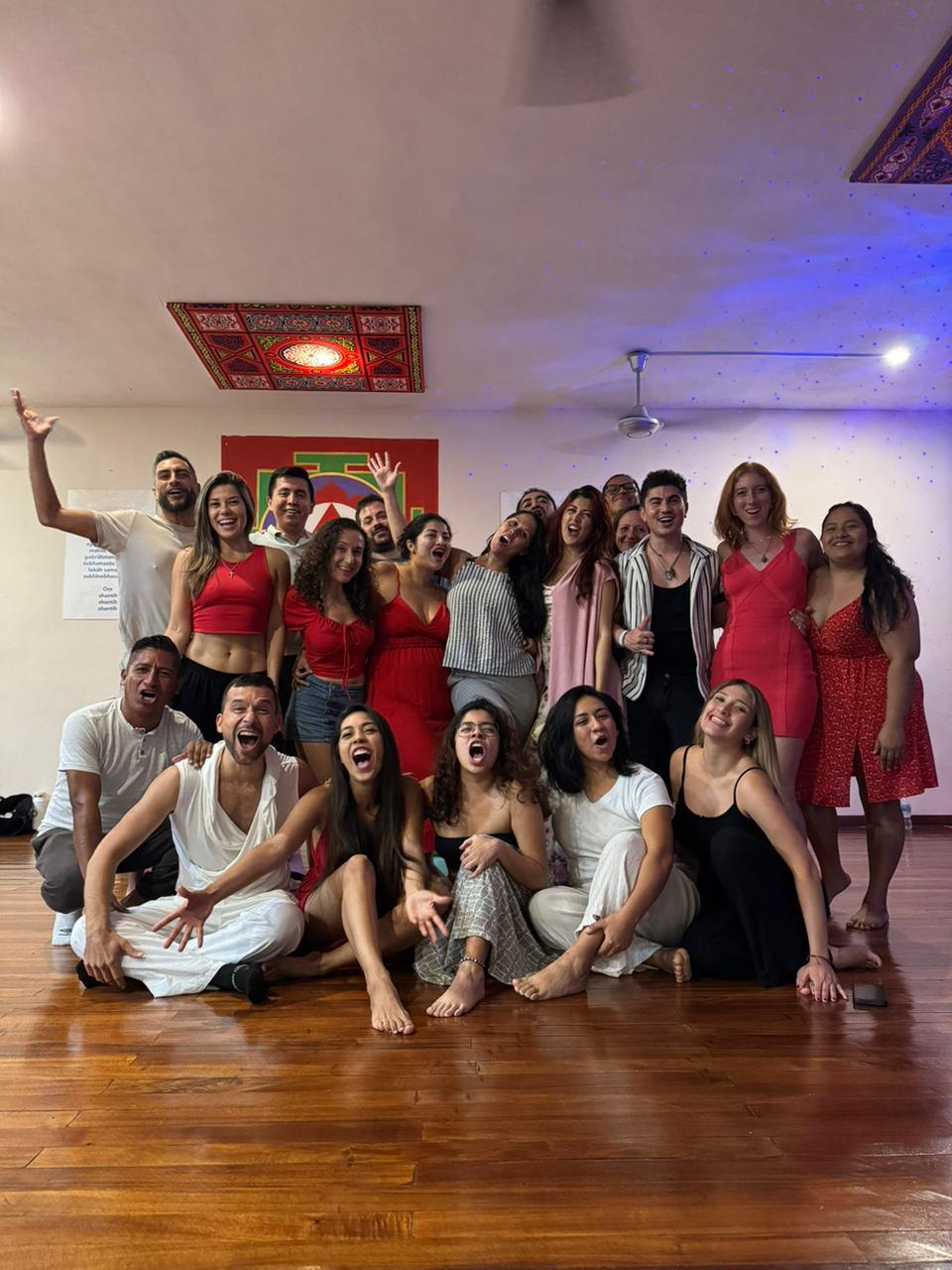 Foto de grupo de hombres y mujeres, varias de rojo, riendo y con la boca abierta en un salón con yantra pintado y piso de madera.