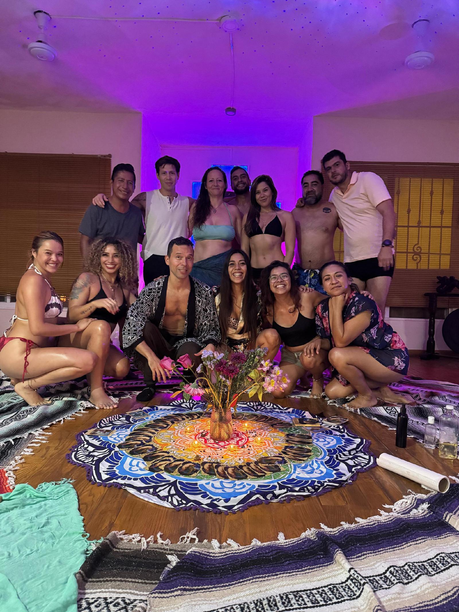 Hombres y mujeres, varios en bikini o sin camisa, posan sonrientes bajo luz violeta tras un altar de flores, velas y cartas sobre un tapete de mandala.