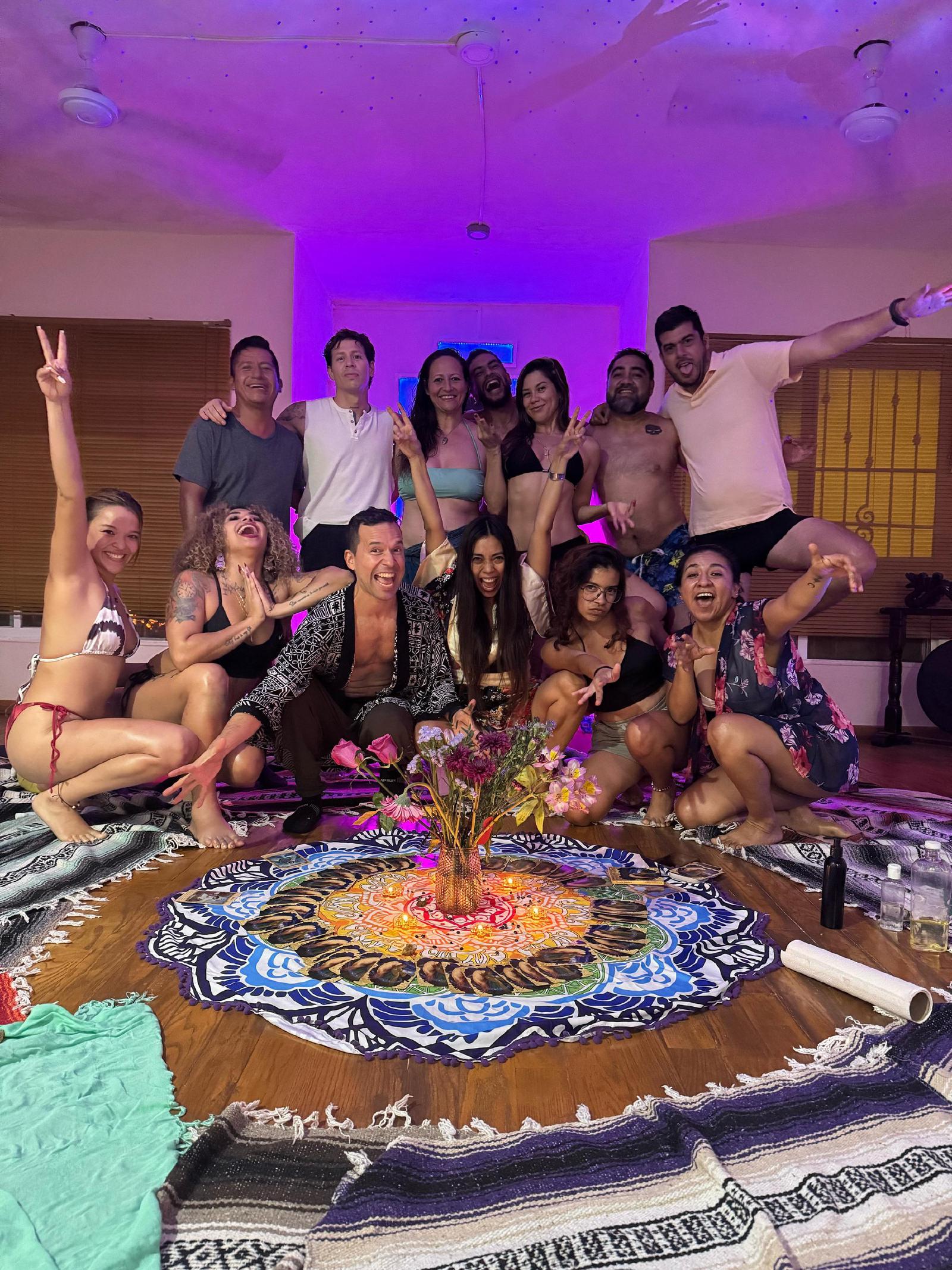 Hombres y mujeres, varios en bikini o sin camisa, posan riendo bajo luz violeta alrededor de un altar de flores, velas y cartas sobre un tapete de mandala.