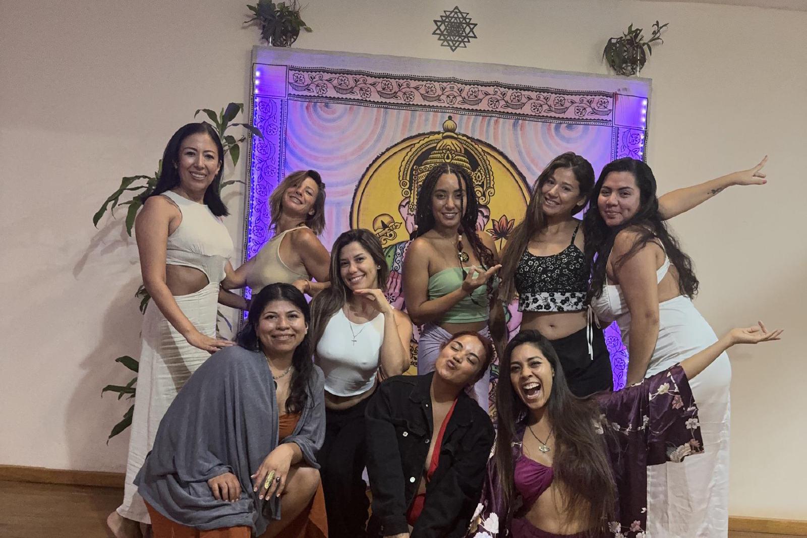 Diez mujeres posan sonrientes frente a un tapiz de Ganesha iluminado en violeta, bajo un techo pintado con la flor de la vida.
