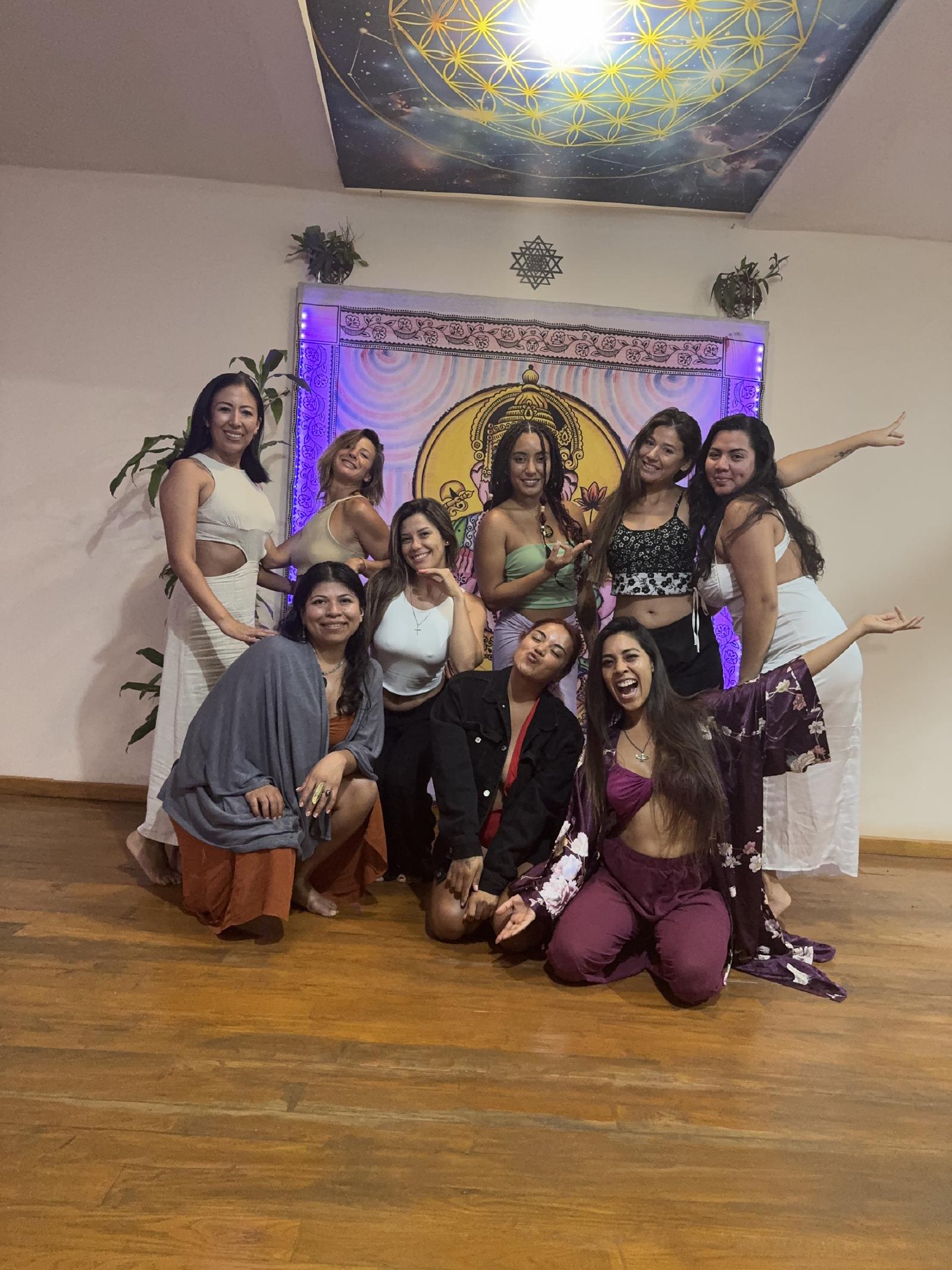 Diez mujeres posan sonrientes frente a un tapiz de Ganesha iluminado en violeta, bajo un techo pintado con la flor de la vida.