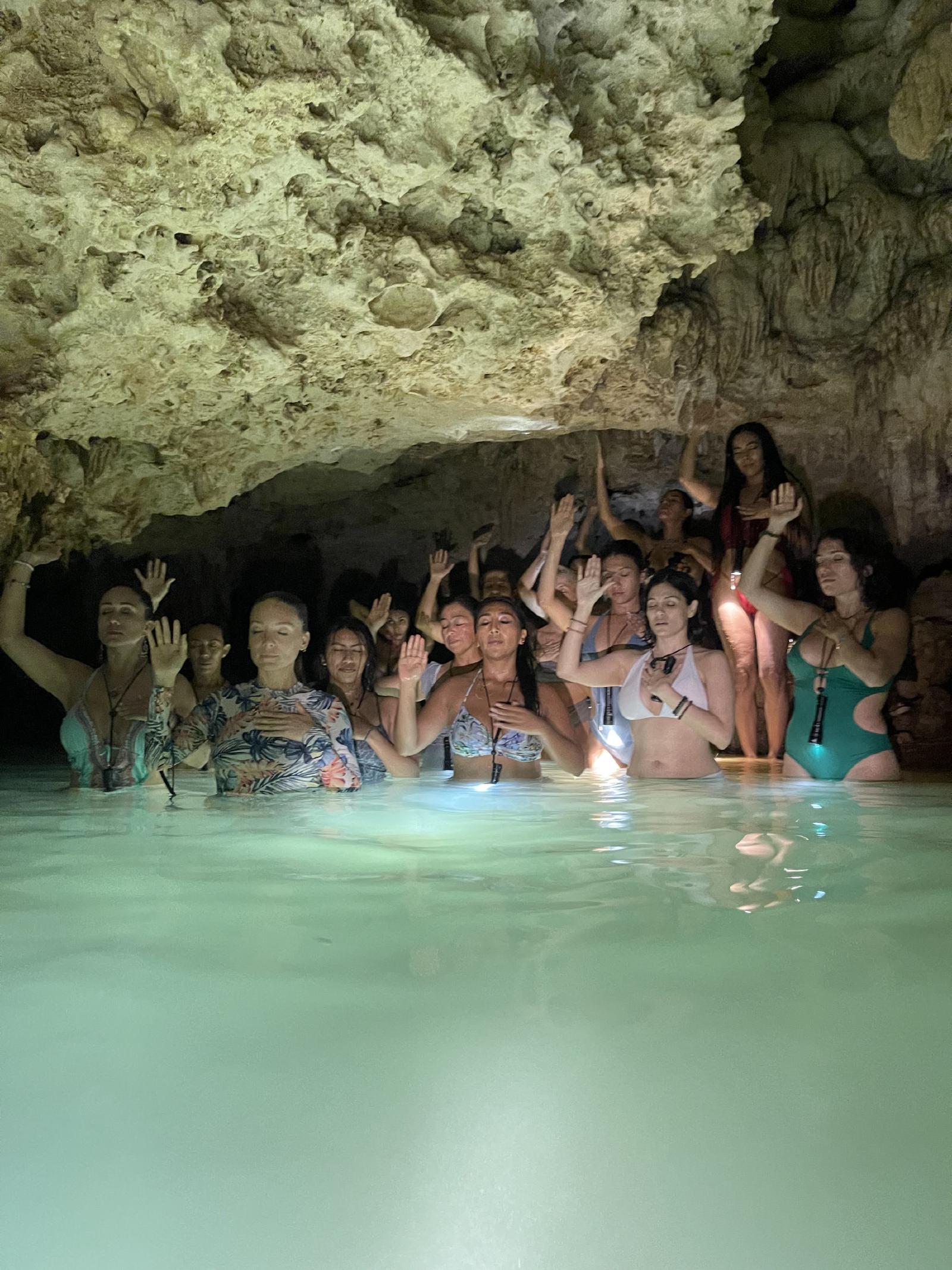 Un grupo de mujeres en traje de baño, de pie en el agua turquesa de un cenote bajo la roca, con los ojos cerrados y una mano en el pecho y otra en alto.