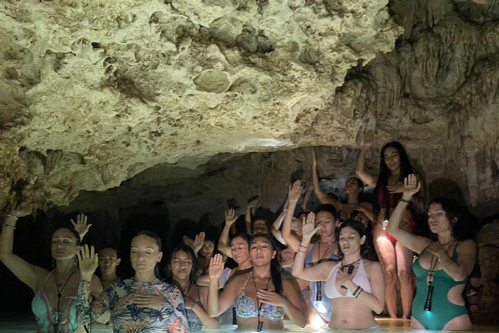 Un grupo de mujeres en traje de baño, de pie en el agua turquesa de un cenote bajo la roca, con los ojos cerrados y una mano en el pecho y otra en alto.