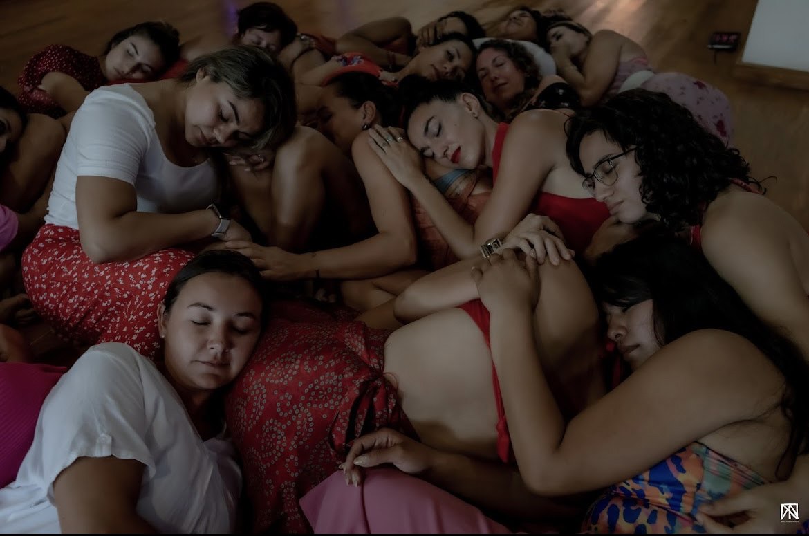Un grupo de mujeres recostadas juntas sobre el suelo, abrazadas y con los ojos cerrados, en ropa blanca, roja y rosa, con luz tenue.