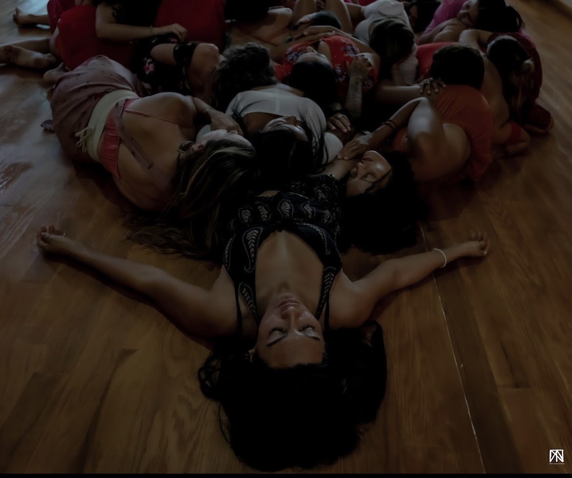 Mujeres recostadas juntas en el suelo de madera, con la cabeza sobre el cuerpo de otra; en primer plano una mujer con los brazos abiertos y los ojos cerrados.