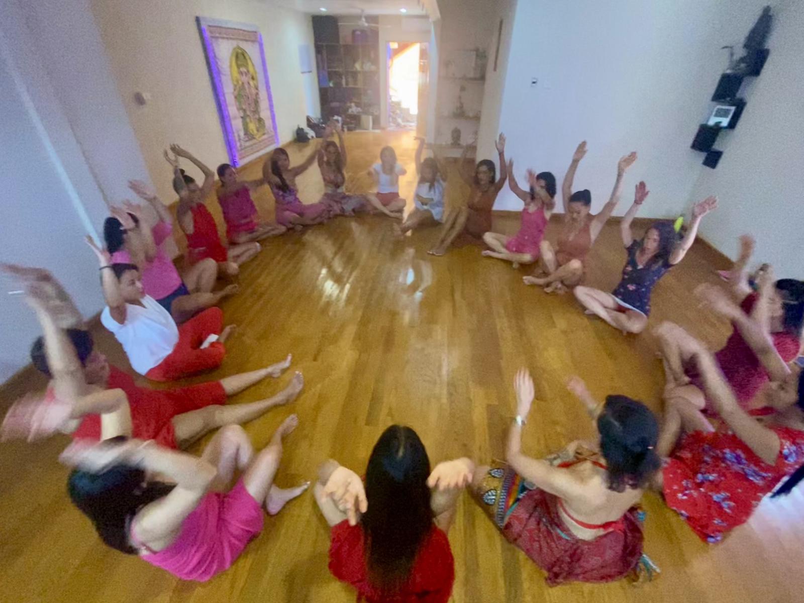 Un círculo de mujeres con ropa roja, rosa y blanca, sentadas en el suelo de madera de una sala, levantan los brazos al mismo tiempo.
