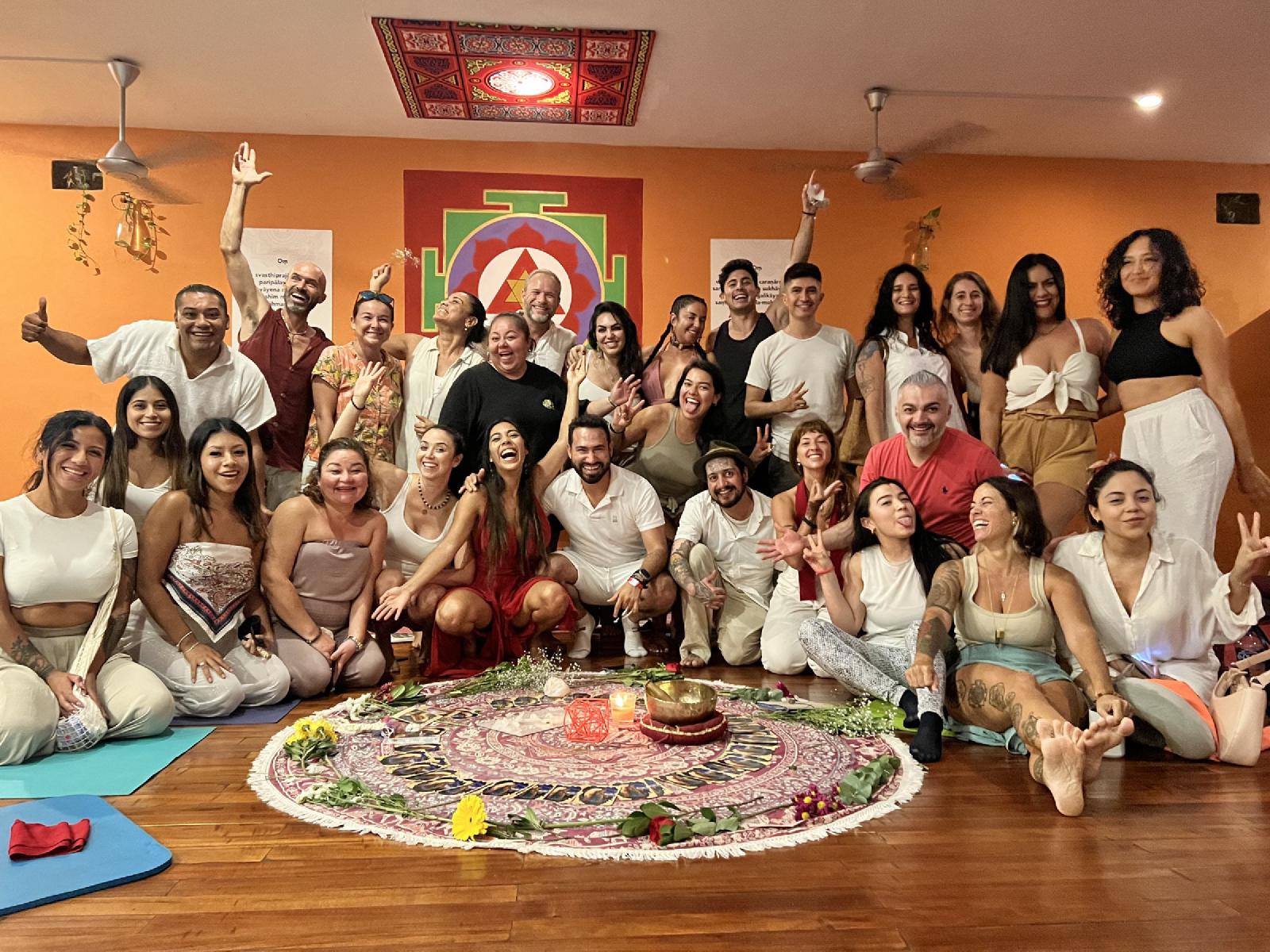 Retrato de grupo alegre de unas treinta personas, casi todas de blanco, sentadas y de pie tras un mandala de flores y velas en una sala naranja con un yantra.