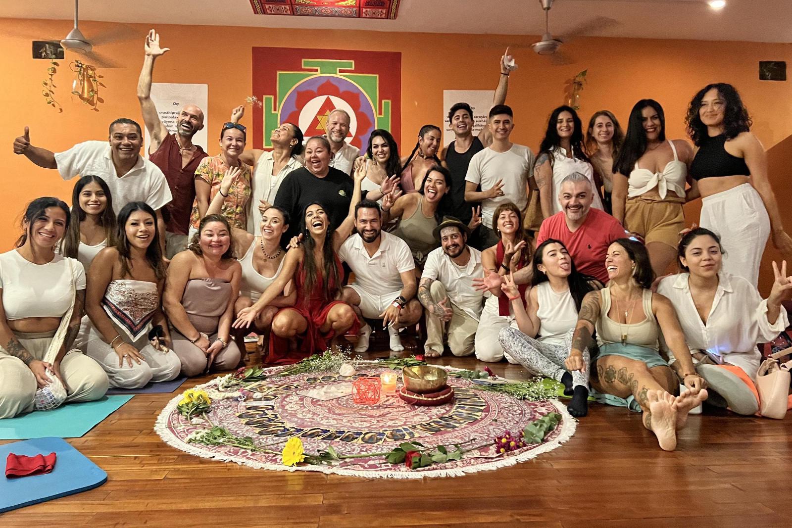 Retrato de grupo alegre de unas treinta personas, casi todas de blanco, sentadas y de pie tras un mandala de flores y velas en una sala naranja con un yantra.
