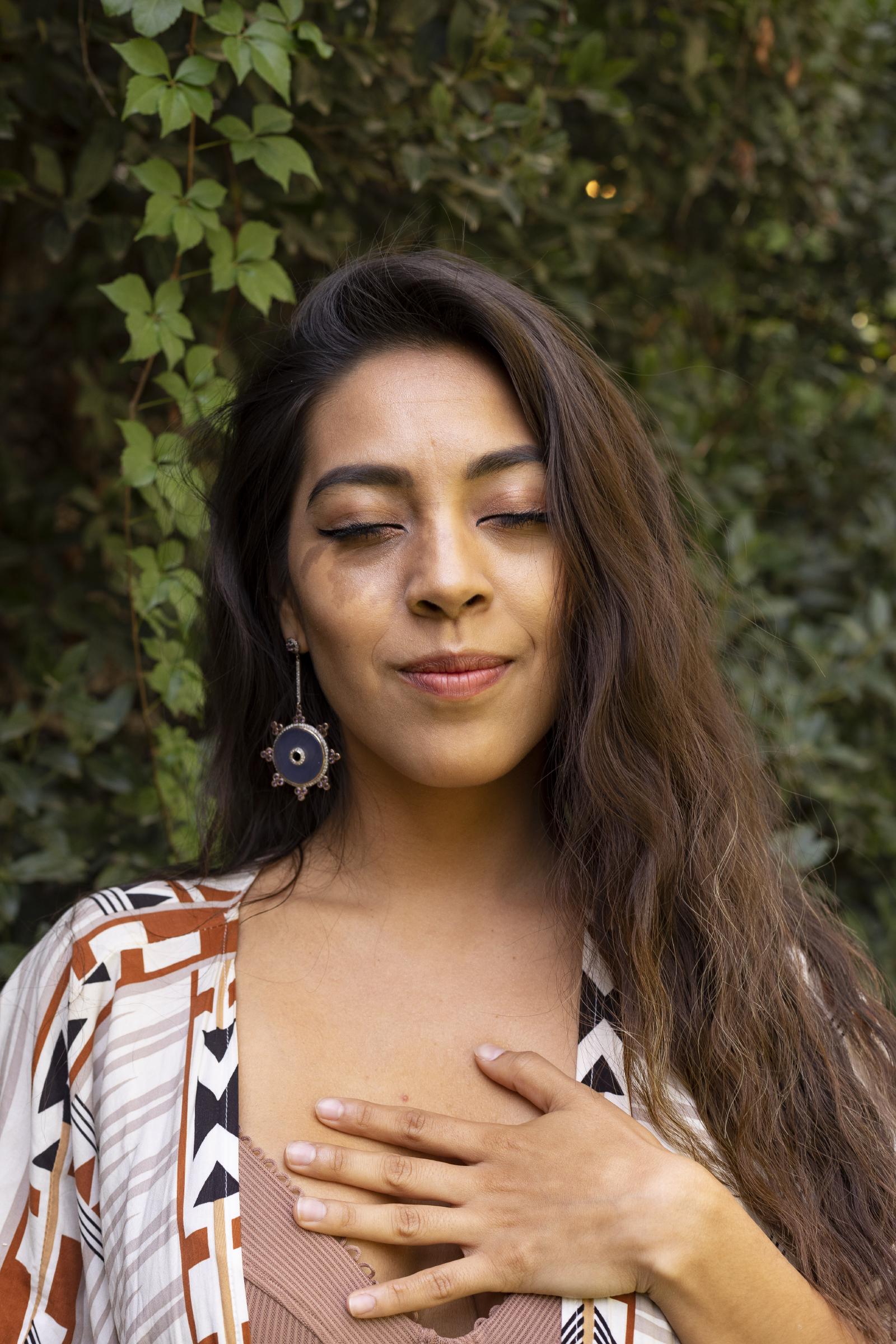 La Dra. Pamela Corona en un jardín, con los ojos cerrados y una sonrisa suave, una mano sobre el corazón