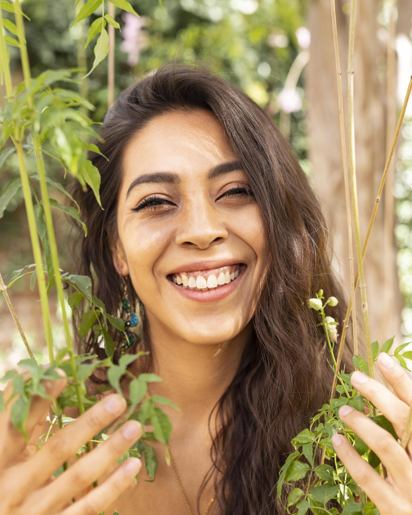 La Dra. Pamela Corona ríe entre ramas verdes en un jardín, con las manos entre las hojas
