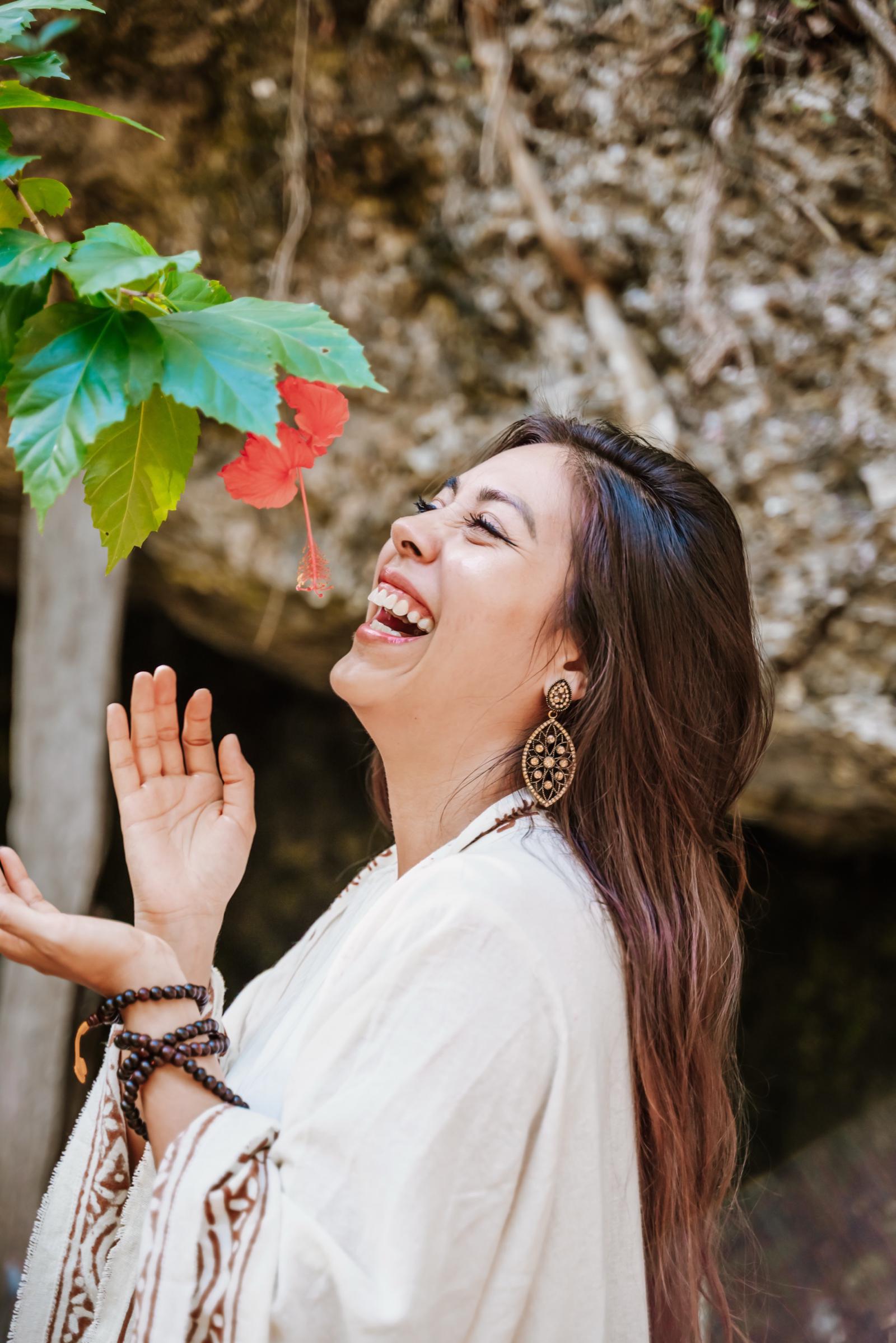 La Dra. Pamela Corona ríe con los ojos cerrados bajo una flor de hibisco roja, frente a una pared de roca