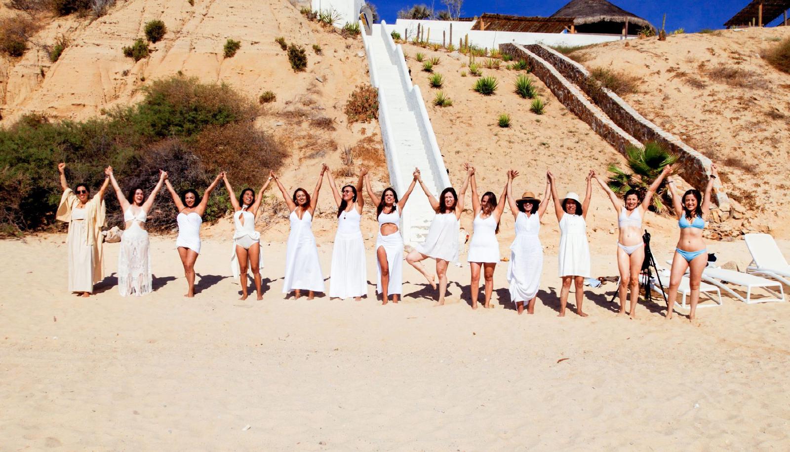 Un círculo de mujeres de blanco en la playa, tomadas de la mano y con los brazos en alto