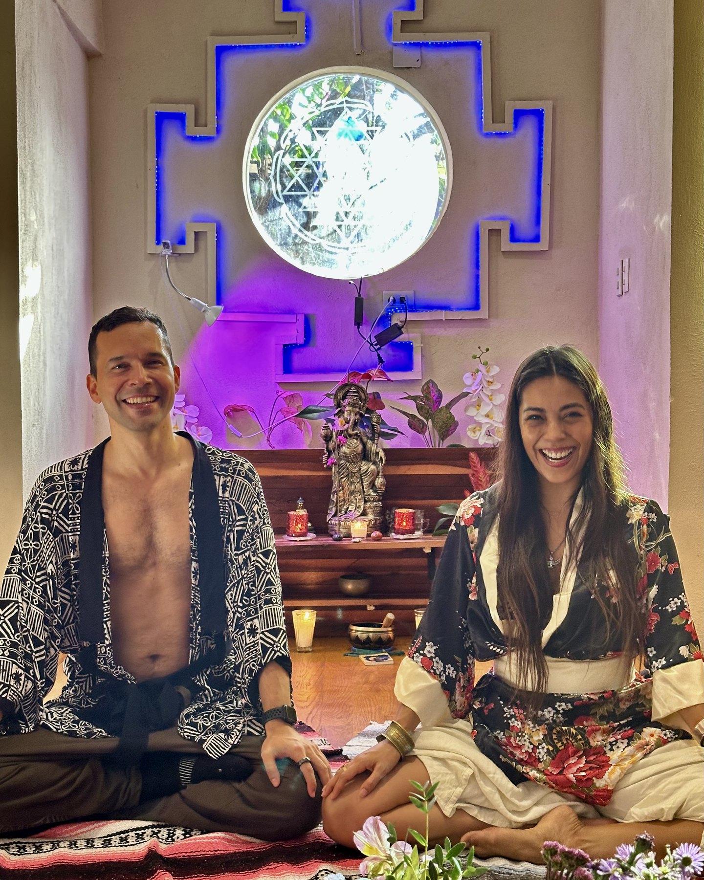 Dos personas sentadas juntas en una sala de práctica bajo un yantra iluminado